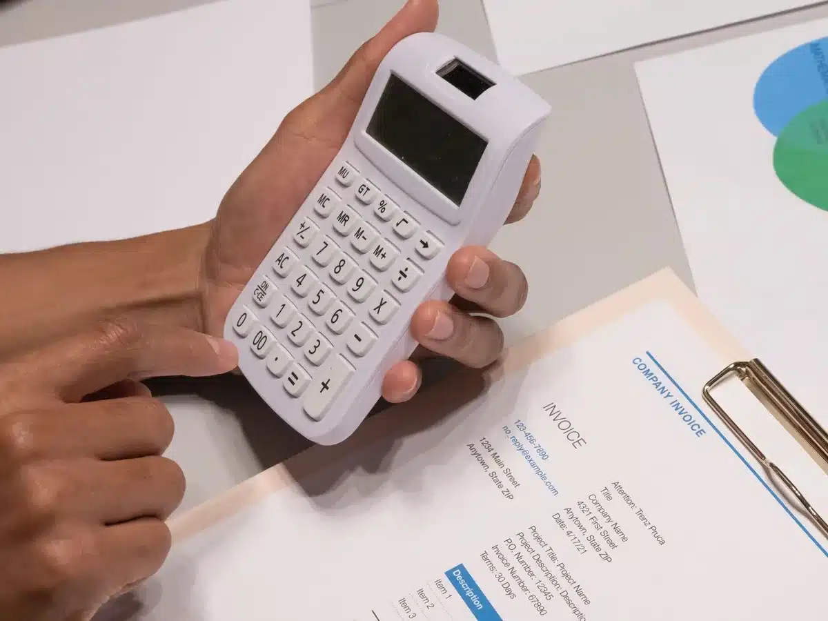 An office desk with hands checking printed invoices and a laptop open to a government website